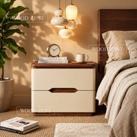 A modern two-drawer nightstand with cream-colored fronts and dark wood accents, styled in a warm, cozy bedroom setting next to a bed with beige bedding and under hanging pendant lights.