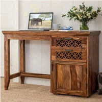 A rustic wooden writing desk with a three-drawer cabinet, featuring decorative lattice cutouts on the middle drawers. The desk is styled with a laptop, mug, glasses, and a vase of flowers against a white paneled wall.